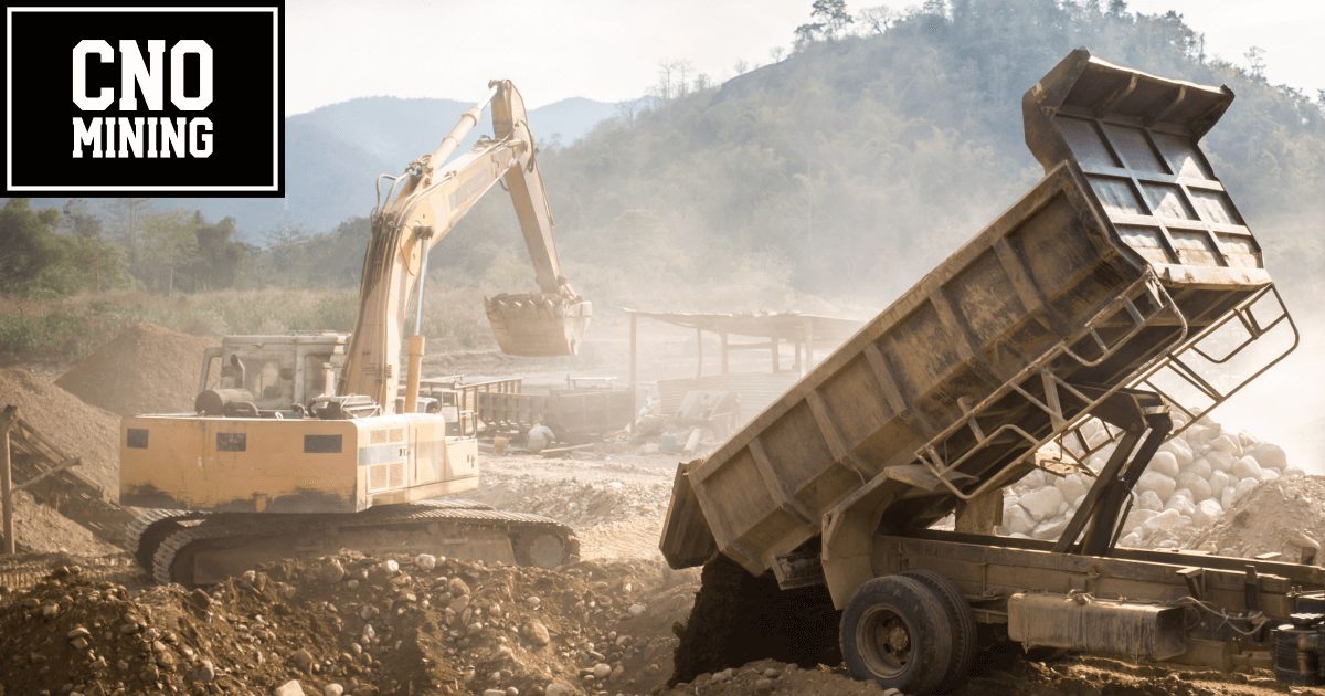 Excavator loading earth into a dump truck at a mining site, with mountains and vegetation in the background, creating a dusty environment